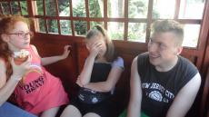 Na zdjęciu widać trójkę nastolatków chroniących się przed słońcem pod altanką. Dziewczynka po lewej stronie ubrana w różową koszulkę zjada kołaczyk z serem.  Dziewczynka na środku w czarnej koszulce zakrywa twarz a chłopiec siedzący po prawej stronie się śmieje.jpg