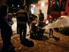 Zdjęcie robione nocą - dwóch chłopców trzyma strażacki wąż z którego leje się woda. W pobliżu widać wóz strażacki na światłach. Chłopcy są asekurowani przez strażaków. .jpg