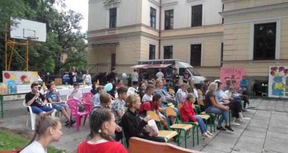 Grupa dzieci i opiekunów siedząca na krzesłach w ogrodzie i oglądająca przedstawienie. Za oglądającymi ustawione są stoły z zimnymi przekąskami. 